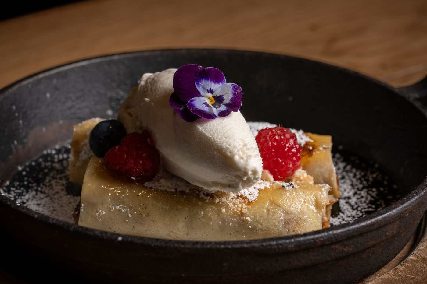 Crepe dulce con crema montada, frutos rojos y flor comestible, servida en plato oscuro sobre fondo negro.